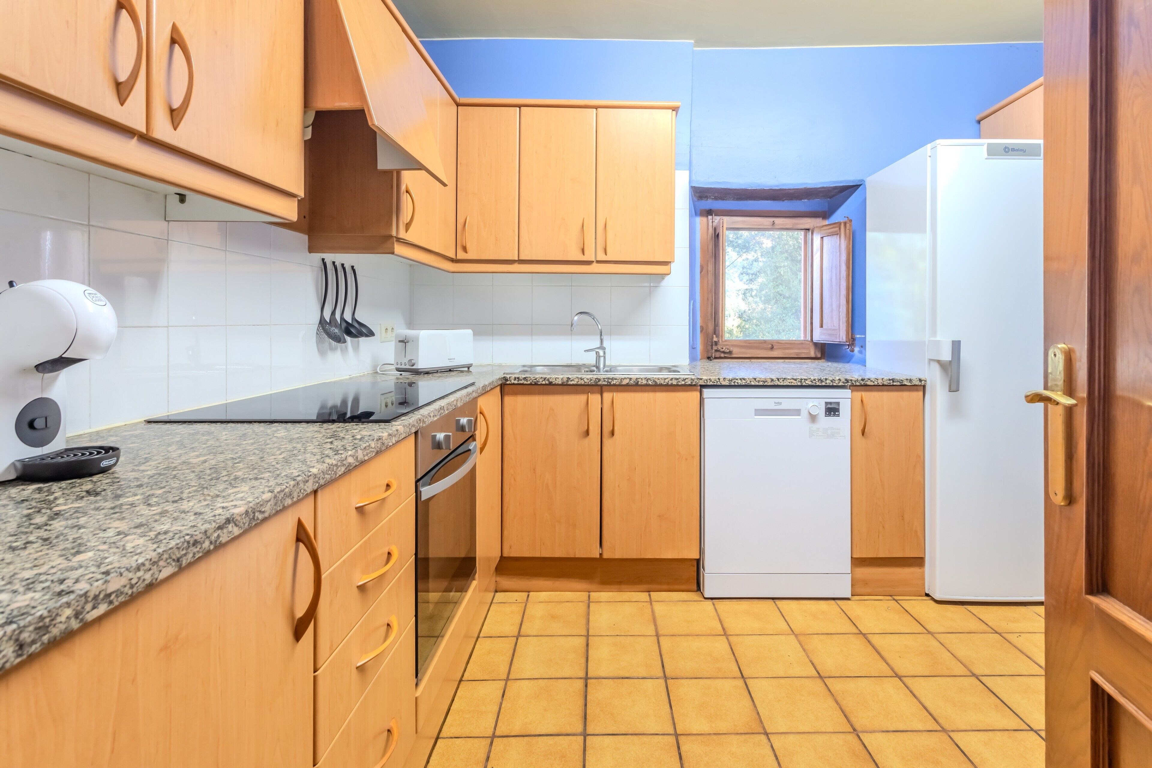 Una cocina con armarios de madera, encimera de granito, lavavajillas y ventana.