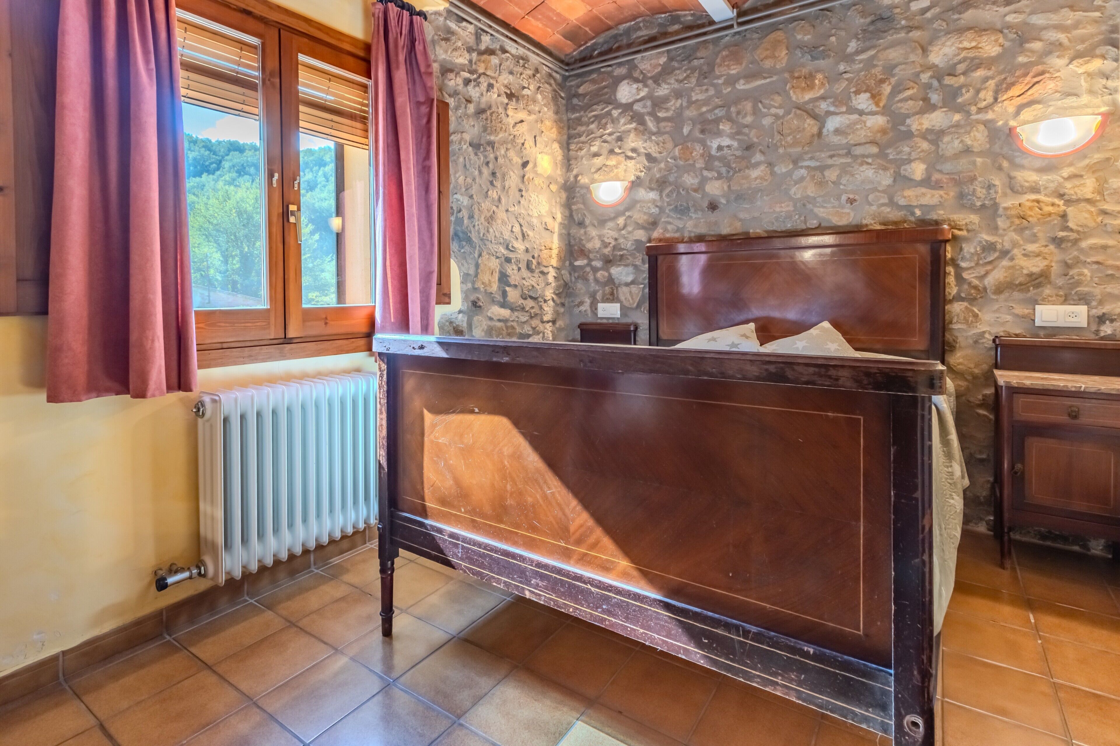 Un dormitorio con pared de piedra, cama de madera, mesita de noche, ventana con cortinas y radiador.