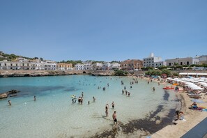 Una playa con gente nadando y tomando el sol, una costa rocosa y edificios al fondo.