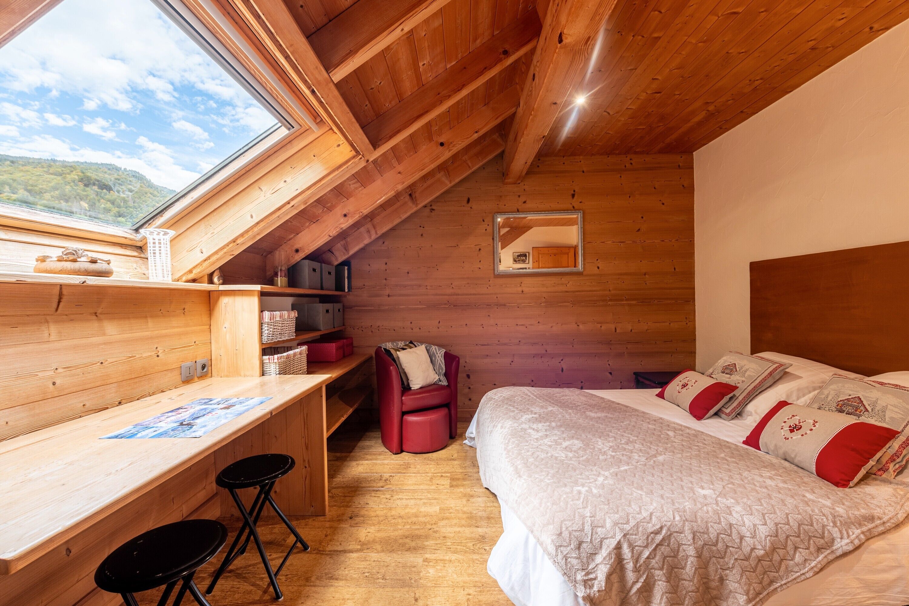Une pièce avec un plafond en pente en bois, un lit avec une literie à motifs, un bureau avec une chaise et une fenêtre donnant sur un paysage naturel.