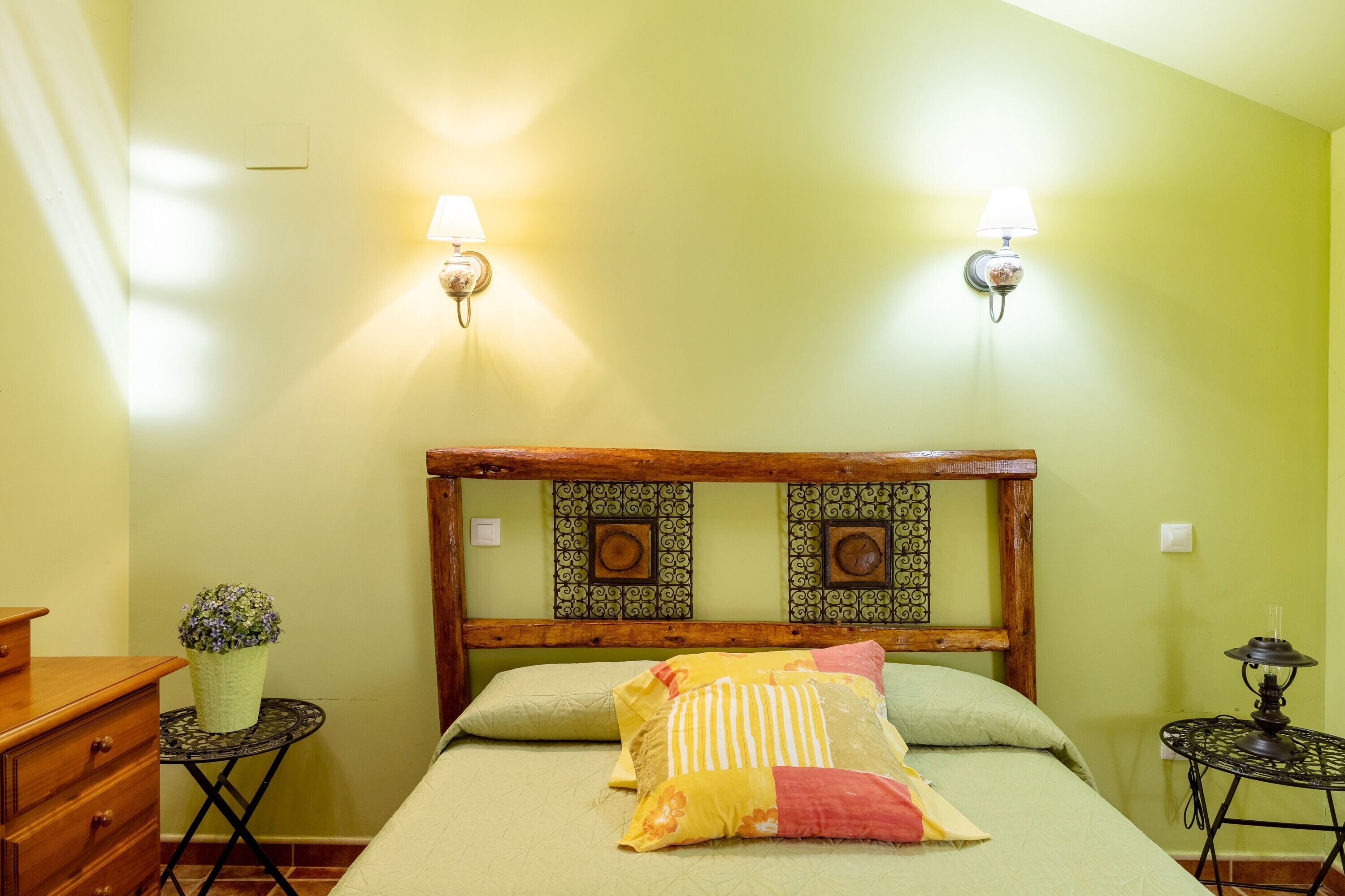 Une chambre avec un lit, deux lampes fixées au mur, une petite table avec une plante et une tête de lit en bois.