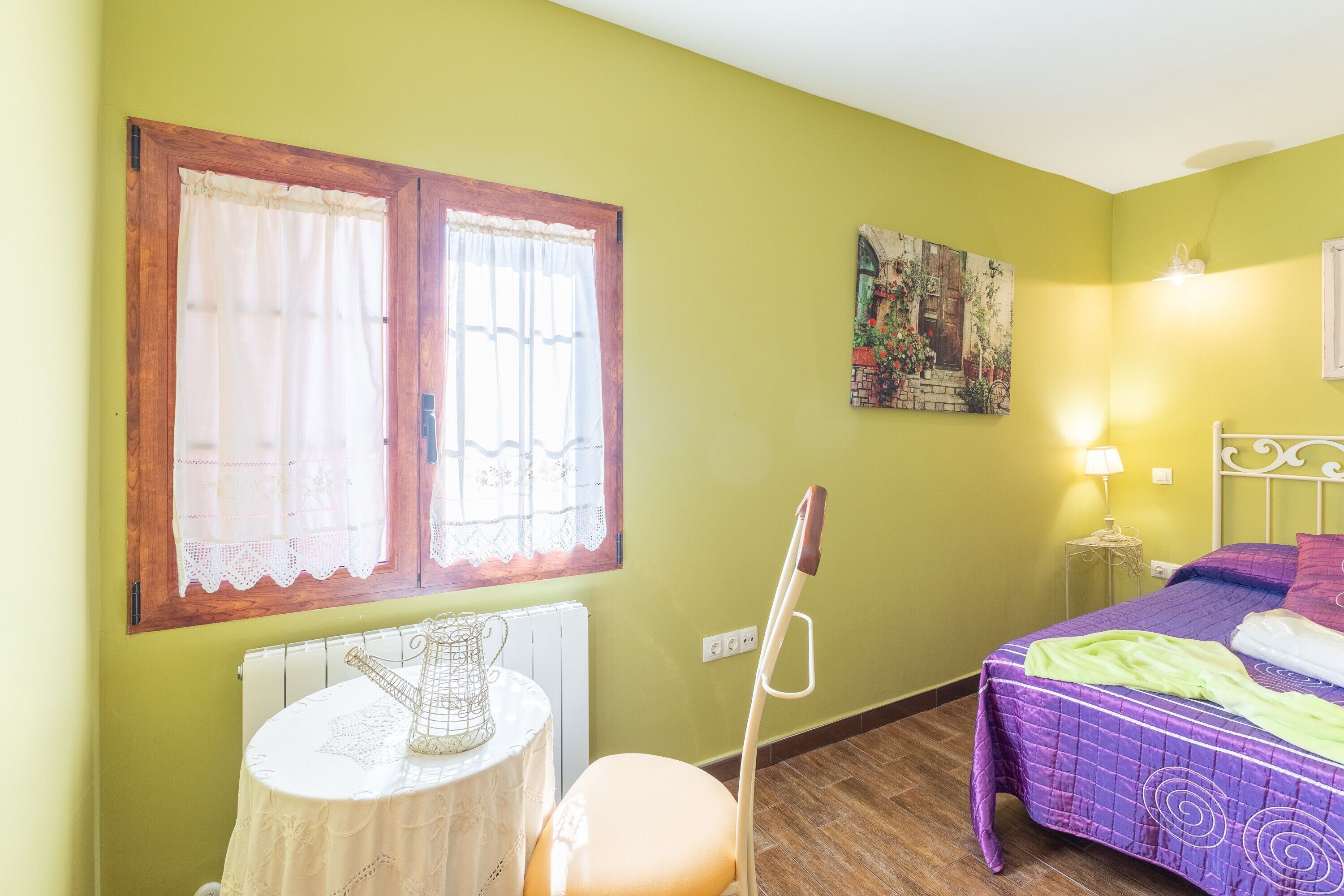 Une pièce avec un lit, une table sur laquelle se trouve une carafe, un radiateur et une fenêtre avec des rideaux.