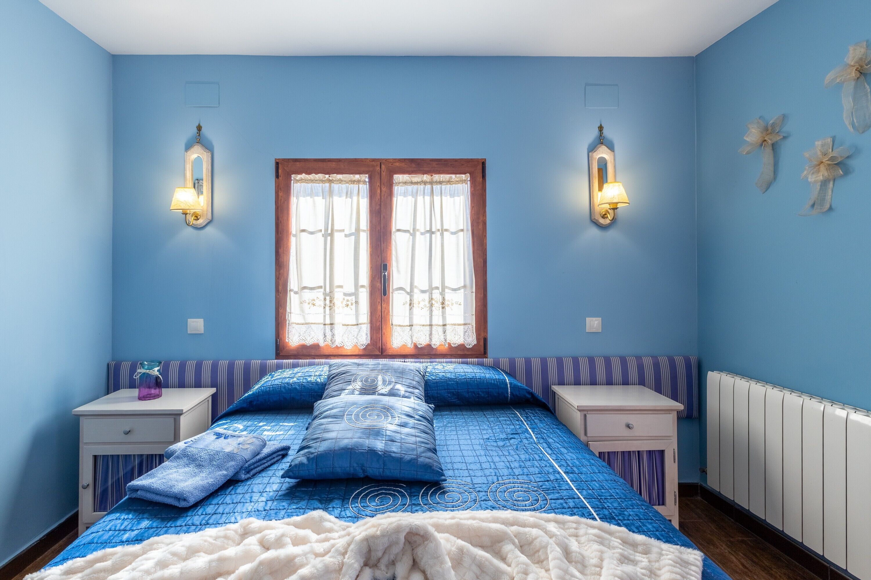 Un dormitorio con cama, dos mesitas de noche, una ventana con cortinas, apliques de pared y un radiador.