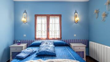 Un dormitorio con cama, dos mesitas de noche, una ventana con cortinas, apliques de pared y un radiador.