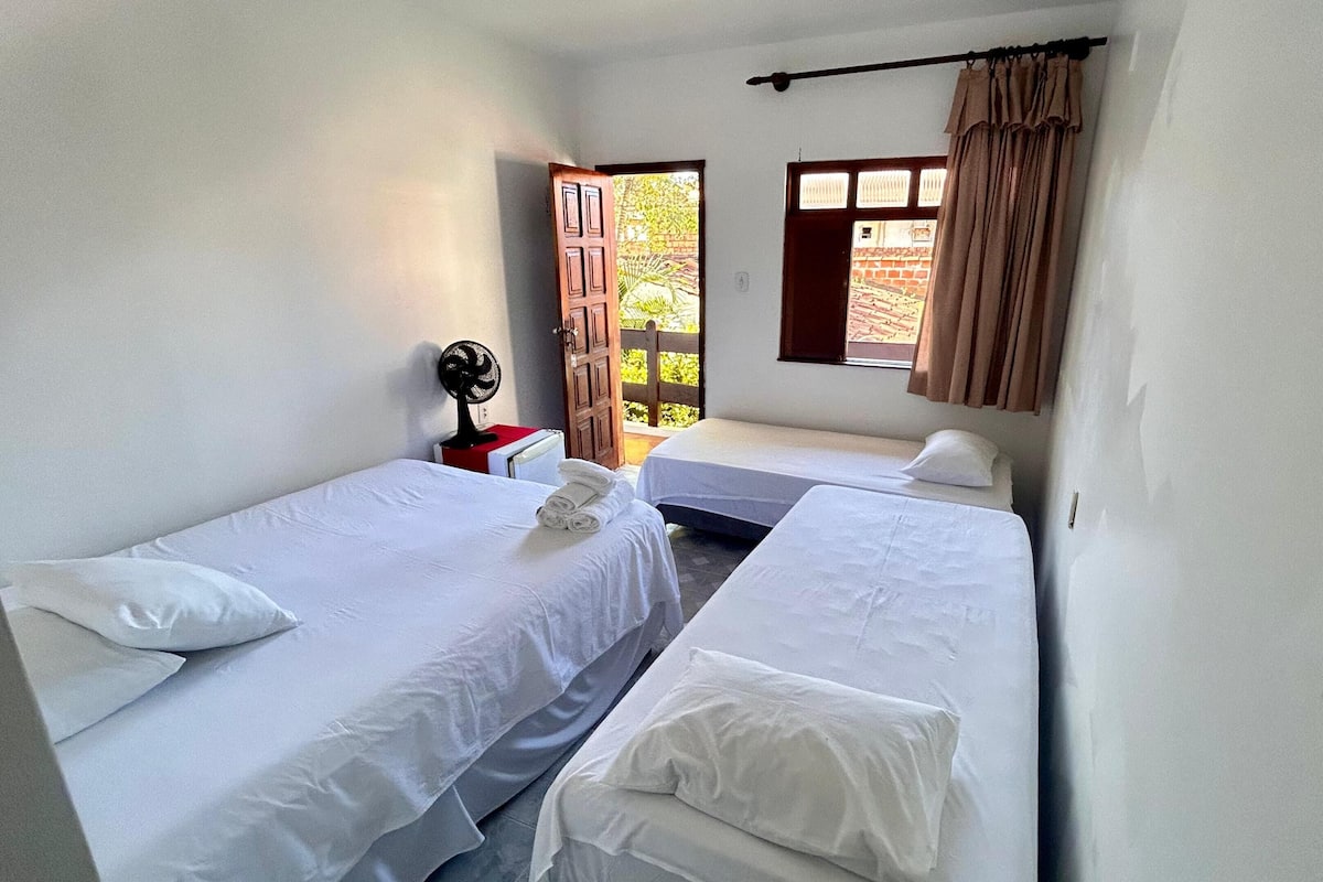 Une chambre avec deux lits, un ventilateur, une chaise rouge et une fenêtre avec des rideaux.