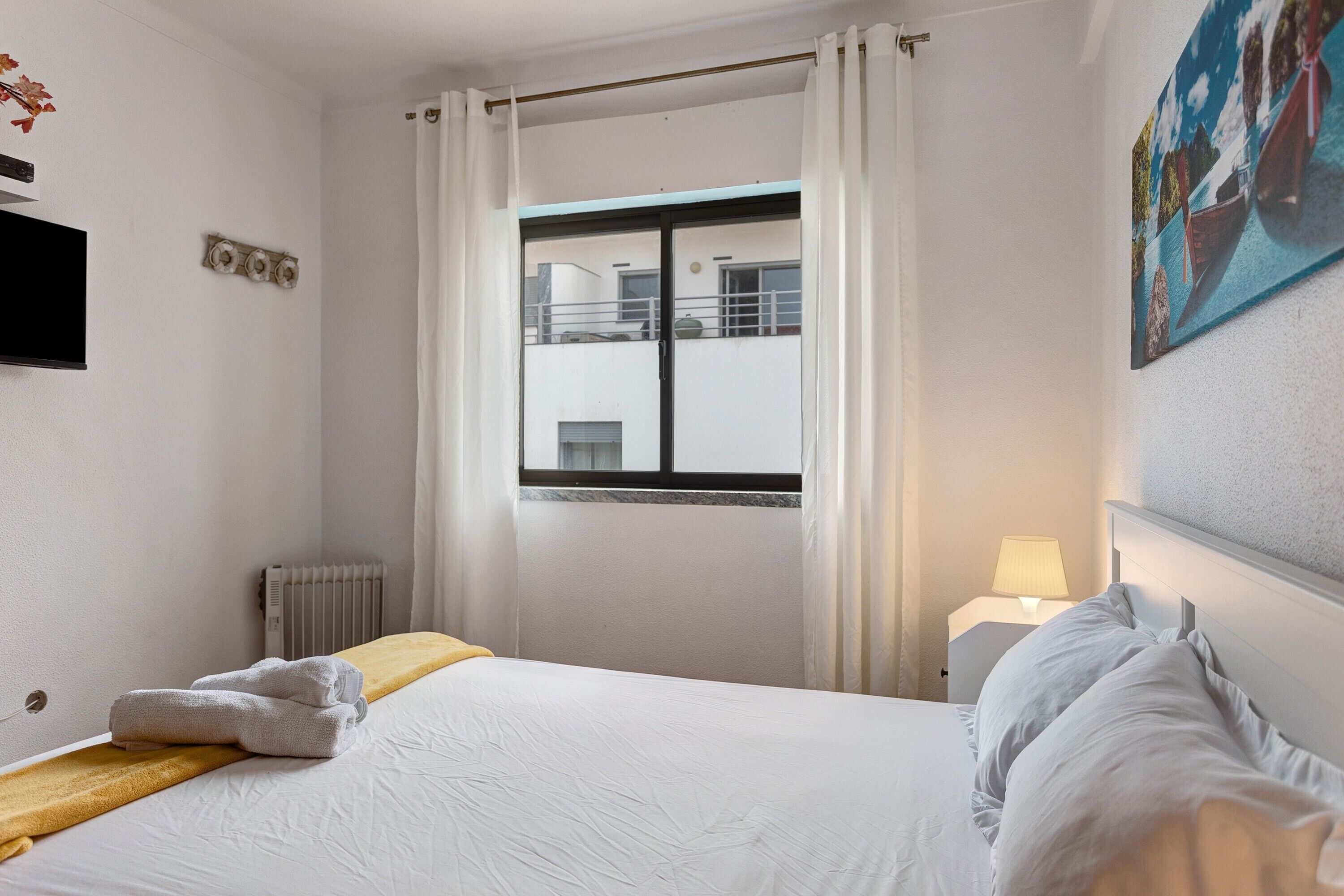 A hotel room with a bed, a window with curtains, a radiator, a TV, and a painting on the wall.