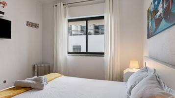 A hotel room with a bed, a window with curtains, a radiator, a TV, and a painting on the wall.