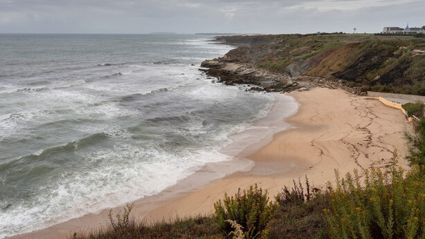 Eine Küstenlandschaft mit Sandstrand, felsigen Klippen und einem Gebäude in der Ferne.