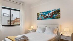 Ein ordentlich bezogenes Bett mit weißen Laken, einer gelben Decke und einem Gemälde einer Küstenlandschaft darüber.