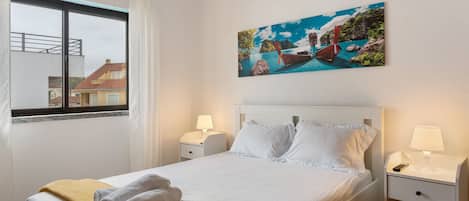 A neatly made bed with white linens, a yellow throw blanket, and a painting of a coastal scene above it.