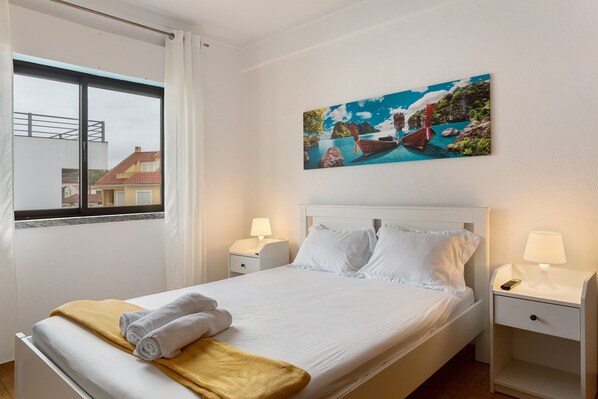 A neatly made bed with white linens, a yellow throw blanket, and a painting of a coastal scene above it.