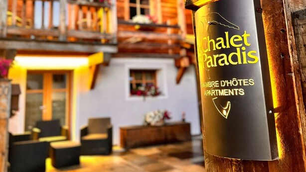 Un chalet en bois doté de balcons, une enseigne indiquant « Chalet Paradis » et une plante en pot.