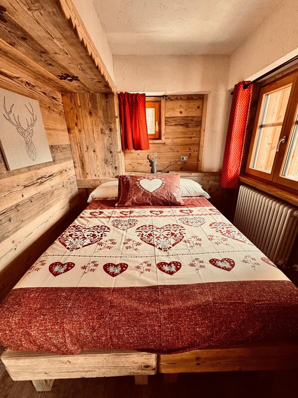 A wooden cabin bedroom with a bed, red curtains, a radiator, and a wall-mounted picture.