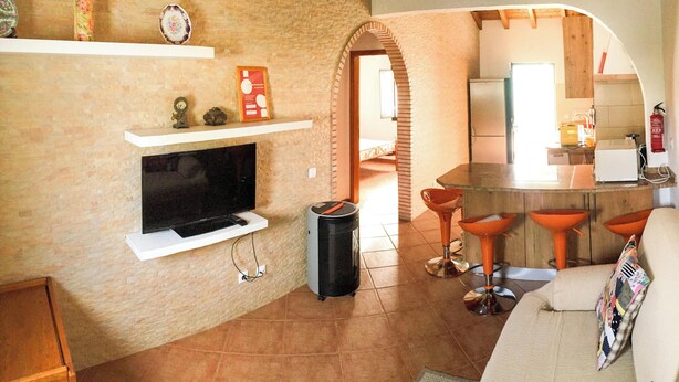 A living room with a TV, a fireplace, a kitchen area, and a dining space with stools. - Cottage 'Casas Da Lagoa - T2 Sul' with Private Garden, Balcony and Wi-Fi (Brescos)