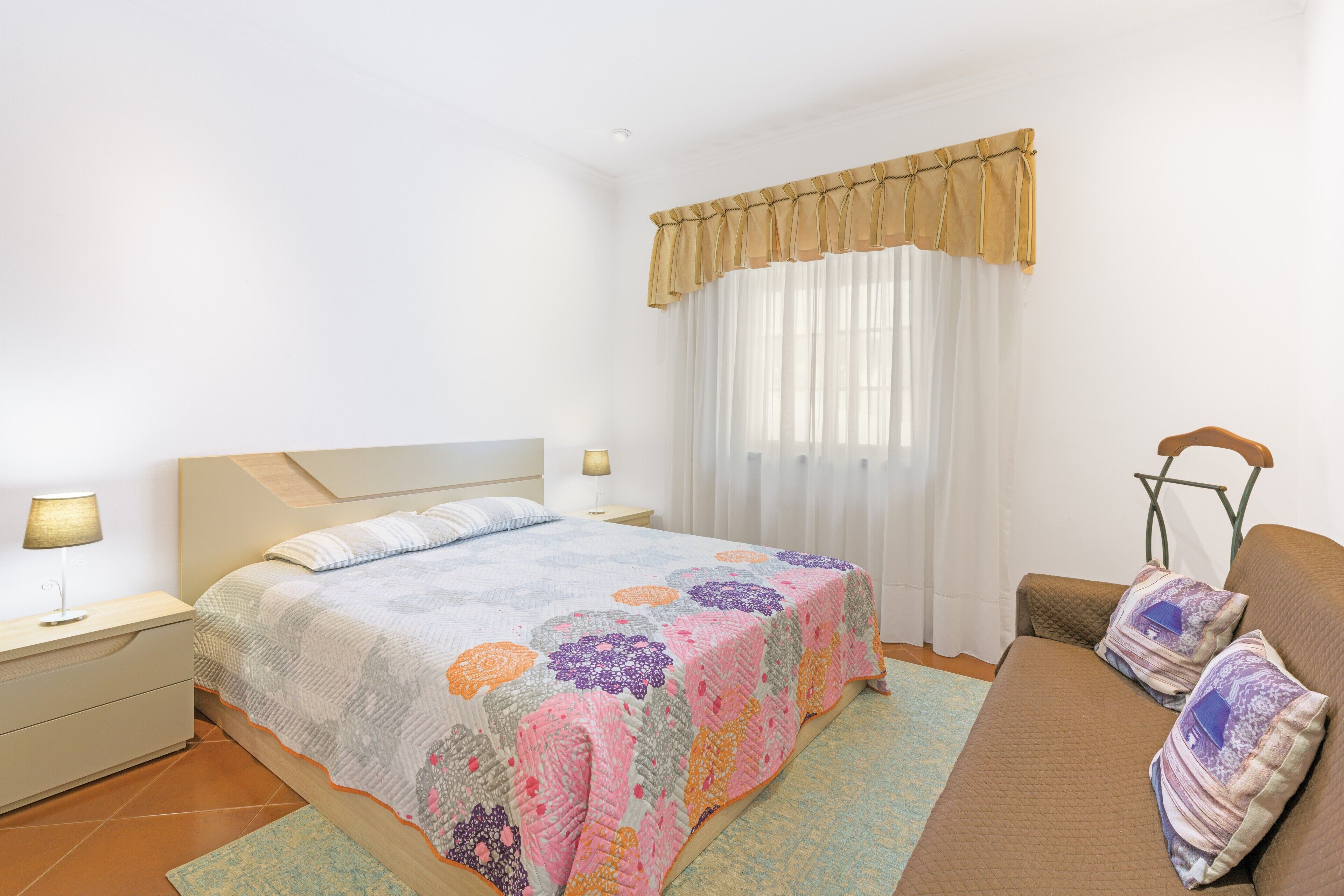 A bedroom with a bed, bedside tables, a nightstand, a sofa, and a window with curtains.