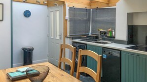 Une cuisine avec une table en bois, des chaises, une horloge, une poubelle et une fenêtre donnant sur un beau paysage.