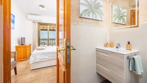 A hotel room with a bed, a wooden dresser, a bathroom with a sink and mirror, and a view through a door. - Apartment 'Vistamar Sa Rapita' with Sea View, Wi-Fi and Air Conditioning (Sa Ràpita)