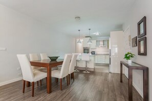 Uma cozinha moderna com armários brancos, uma sala de jantar com mesa e cadeiras de madeira, e um corredor que dá para uma área de estar.