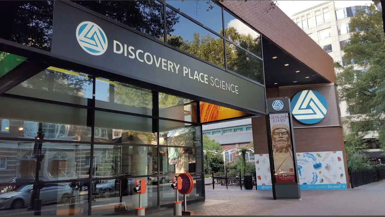 The exterior of Discovery Place Science with large glass windows, a prominent entrance, and a logo on the facade.
