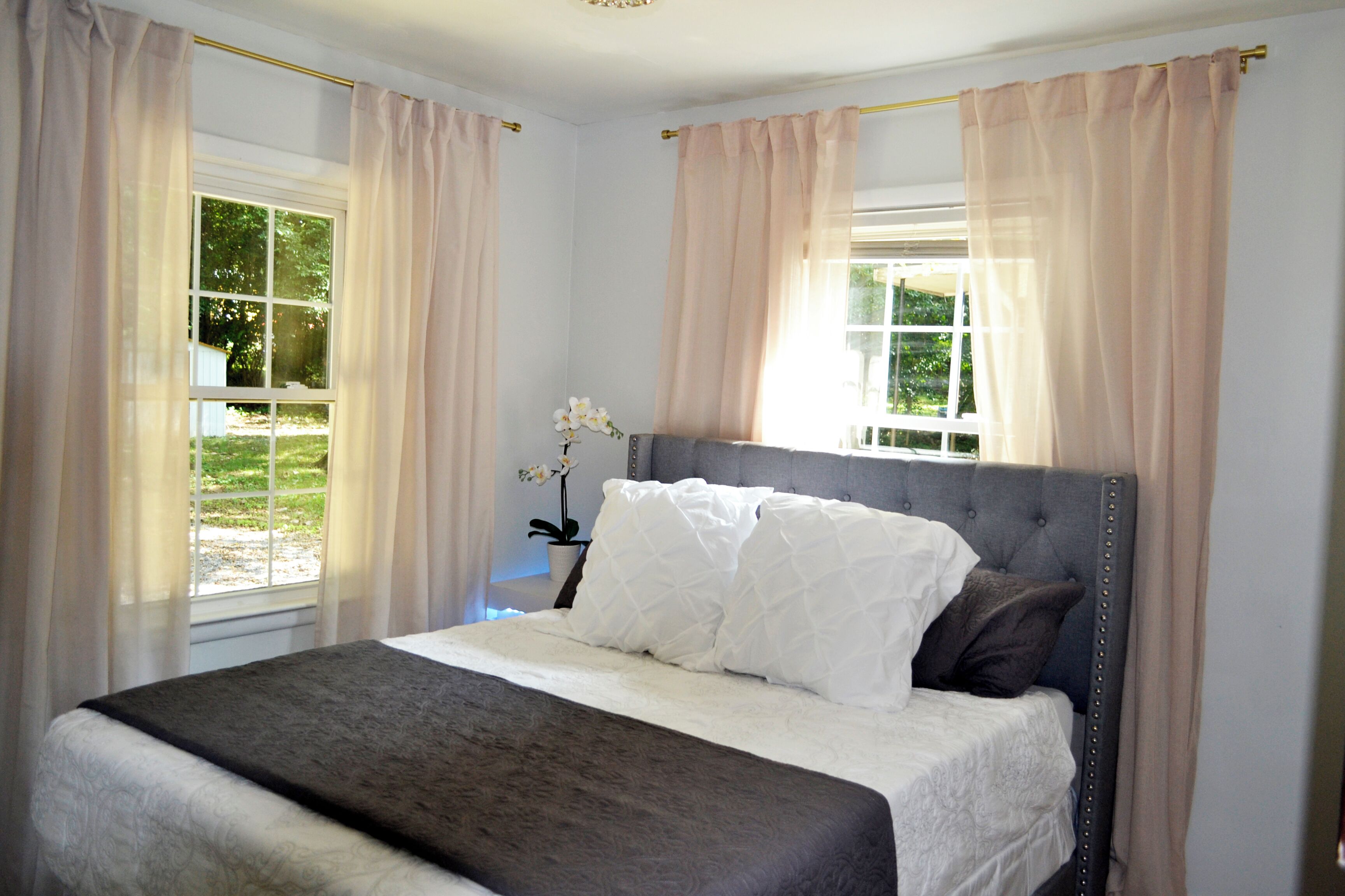 Una cama bien hecha con cabecera gris y ropa de cama blanca, una planta en maceta sobre la mesita de noche y cortinas traslúcidas en las ventanas.