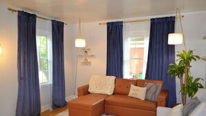 Ein Wohnzimmer mit einem braunen Ecksofa, einem weißen Sessel und einem Couchtisch. Zwei Fenster mit blauen Vorhängen, eine Stehlampe und ein Topf mit Pflanze sind ebenfalls zu sehen.