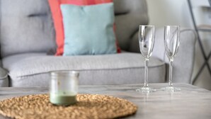 Una sala de estar con un sofá gris, una almohada azul y roja, dos vasos transparentes y un pequeño recipiente de vidrio sobre un posavasos tejido.