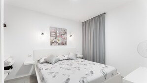 Un dormitorio con cama, mesita de noche, lámpara de pared, cortina y una obra de arte en la pared.