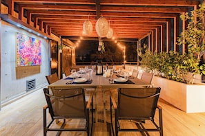 A dining area with a long table set for four, wooden beams on the ceiling, and a colorful abstract painting on the wall.