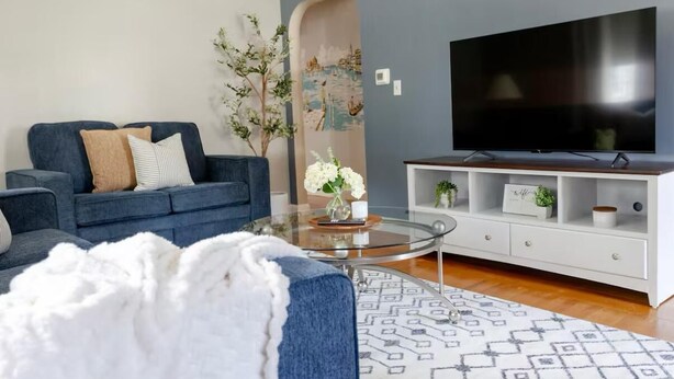 Een moderne woonkamer met een blauwe bank, een glazen salontafel, een witte tv-meubel en een grote flatscreen televisie.