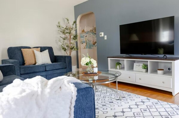 Una sala de estar moderna con un sofá azul, una mesa de centro de vidrio, un mueble blanco para entretenimiento y un televisor de pantalla plana grande.