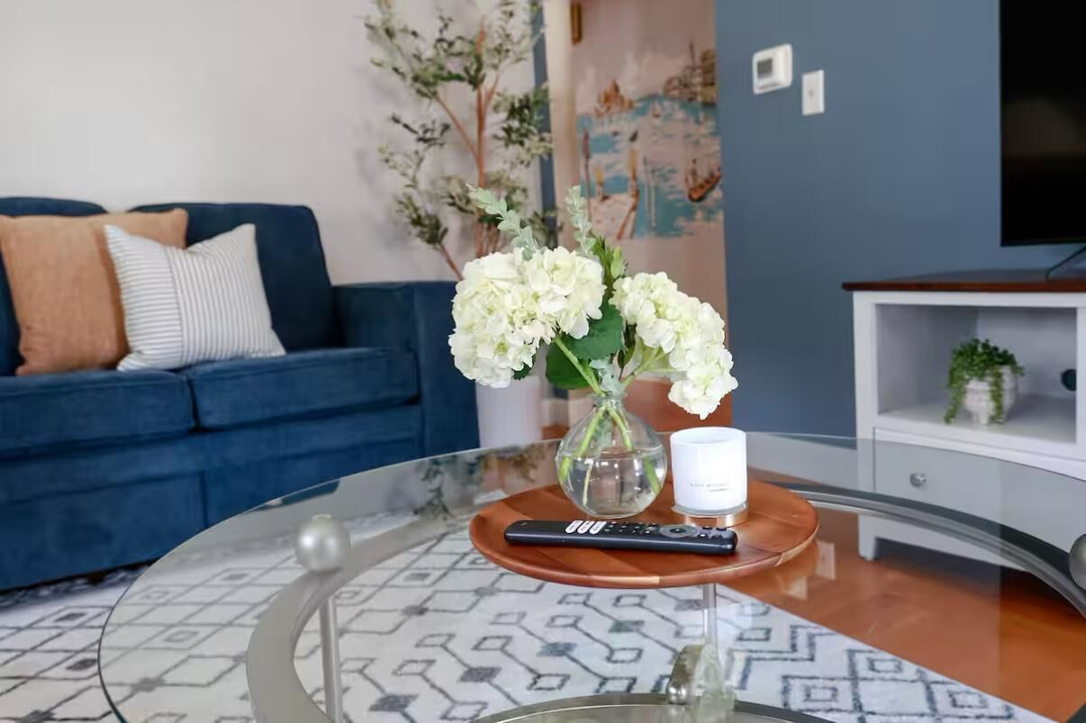 Un saló amb un sofà blau, una taula de centre de vidre, un vas amb flors blanques, un comandament a distància i una tassa blanca.