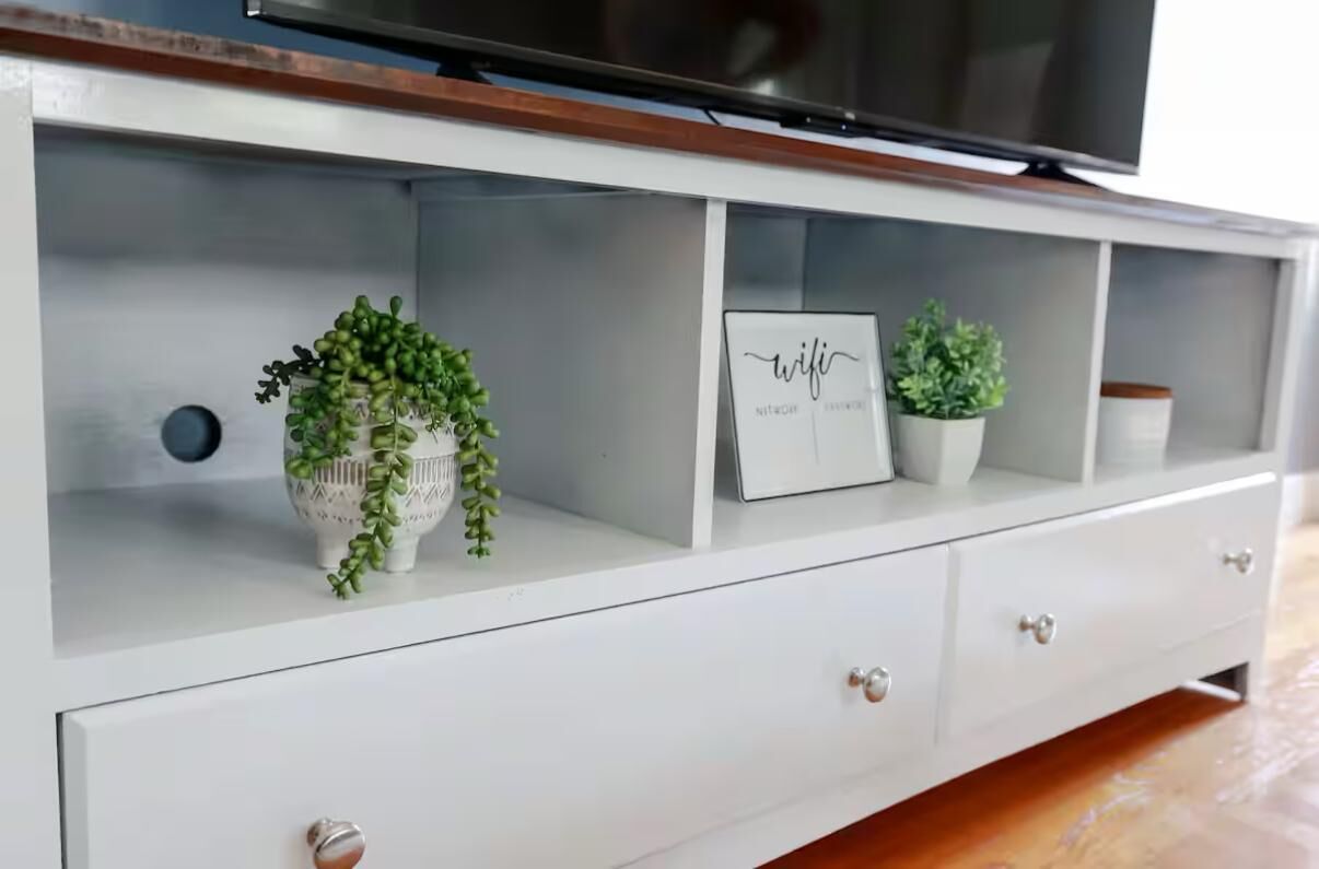 Un meuble blanc pour télévision, avec un téléviseur à écran plat posé dessus, trois tiroirs et deux plantes en pot.