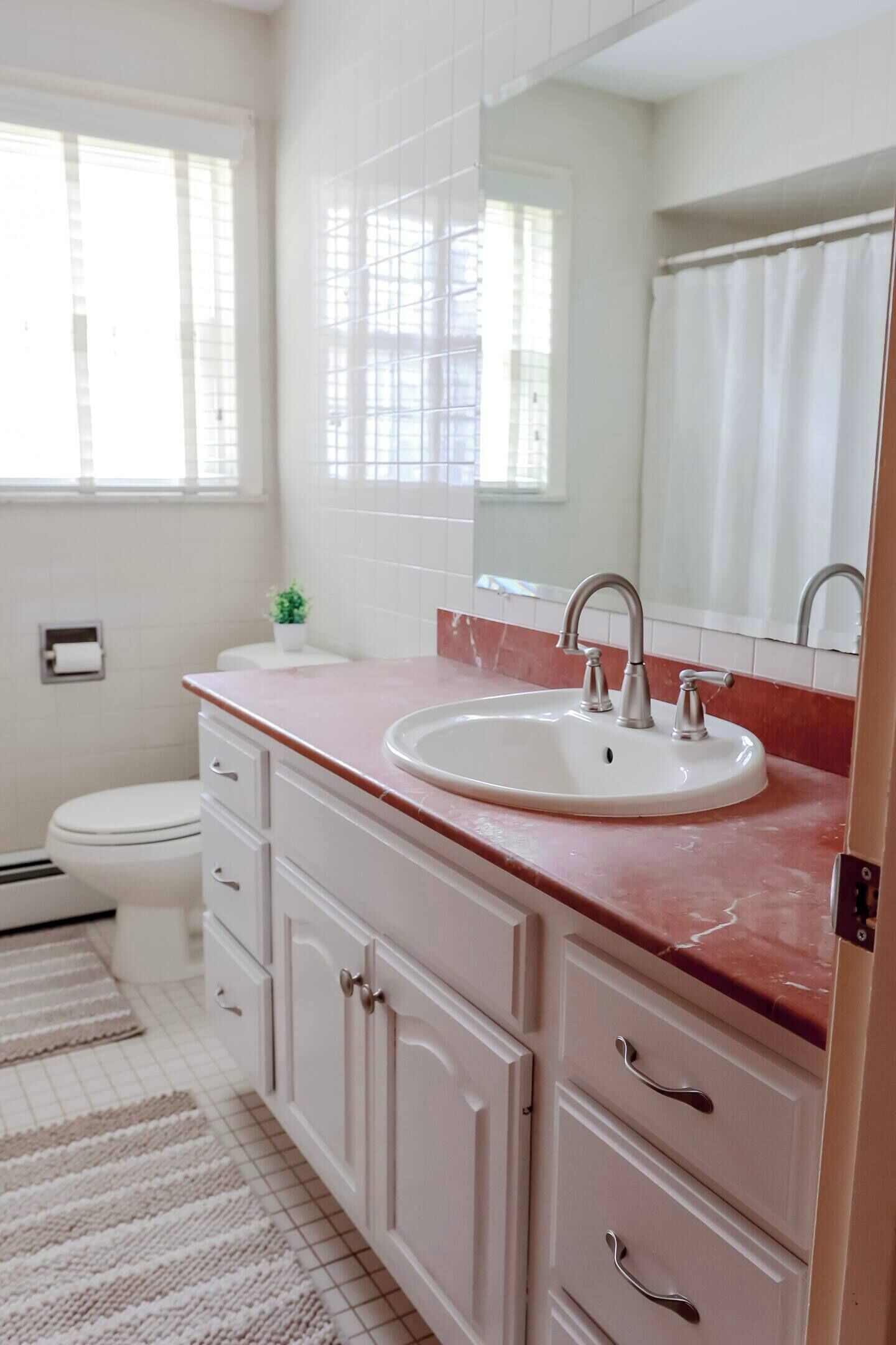 Un baño con un lavabo blanco, un espejo y un inodoro. Hay una ventana con persianas y una planta pequeña en el mostrador.