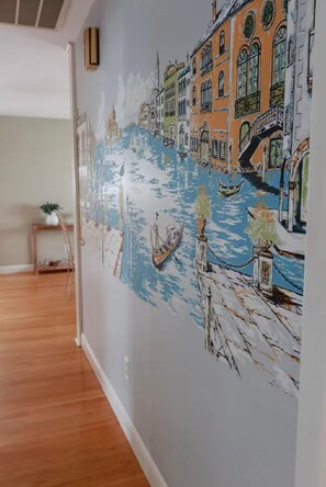 Un pasillo con un mural que muestra una escena de canal, suelo de madera y una pequeña mesa con una planta.