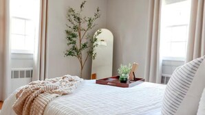 Een netjes opgemaakt bed met een witte deken en een beige plaid, een nachtkastje met een dienblad waarop een plantje en een boek liggen, en een grote spiegel aan de muur.