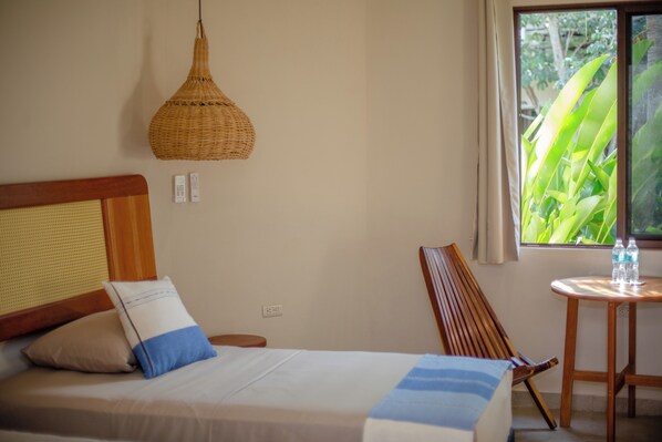 Una cama bien hecha con almohadas, una lámpara colgante de tejido, una silla de madera, una mesita con botellas de agua y una ventana con vistas a la vegetación.