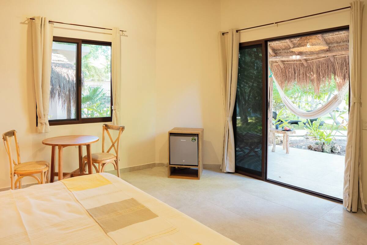 Ein Hotelzimmer mit Bett, einem kleinen Tisch, zwei Stühlen, einer Minibar und einer Glastür zum Außenbereich mit Hängematte und Pflanzen.