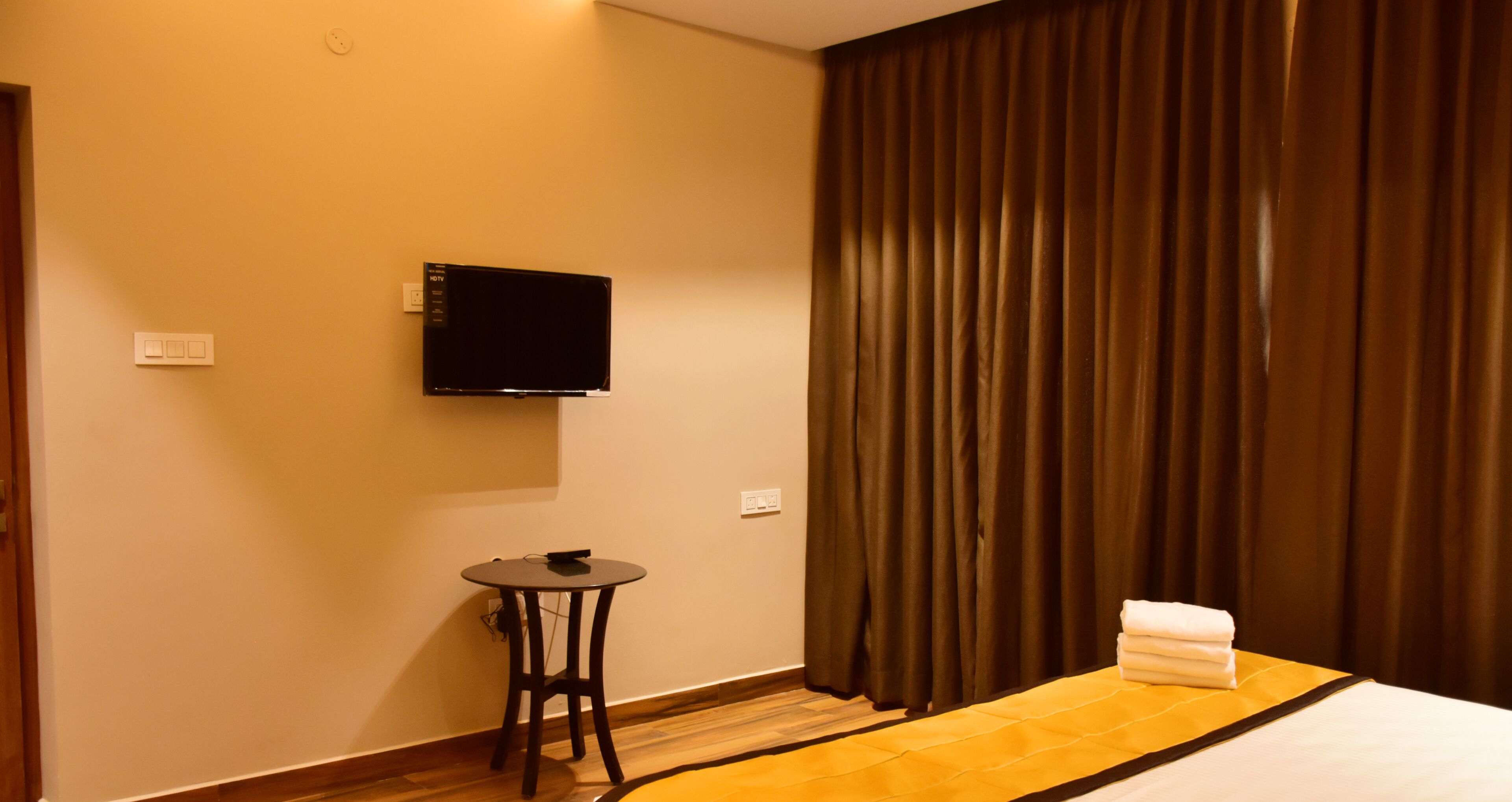 A hotel room with a bed, a flat-screen TV mounted on the wall, a small round table, and a curtain.