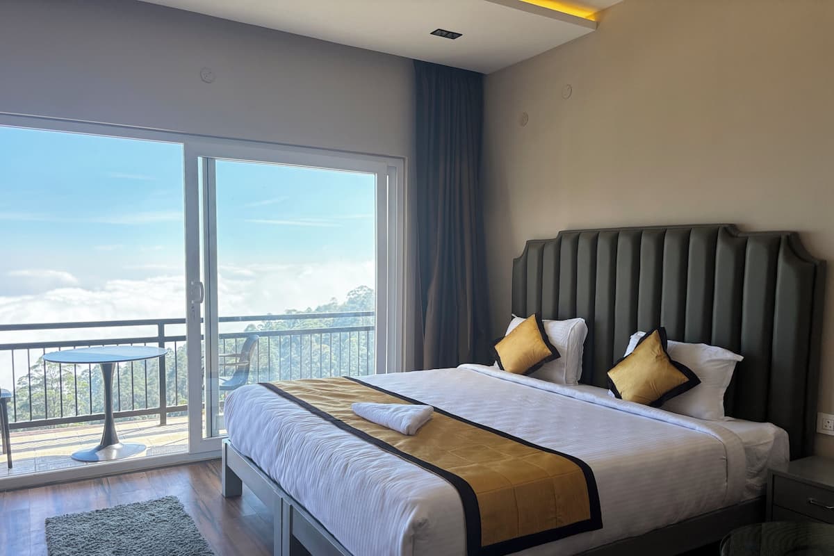 A modern hotel room with a large bed, a glass door leading to a balcony with a view of the clouds and trees, and a small table by the window.