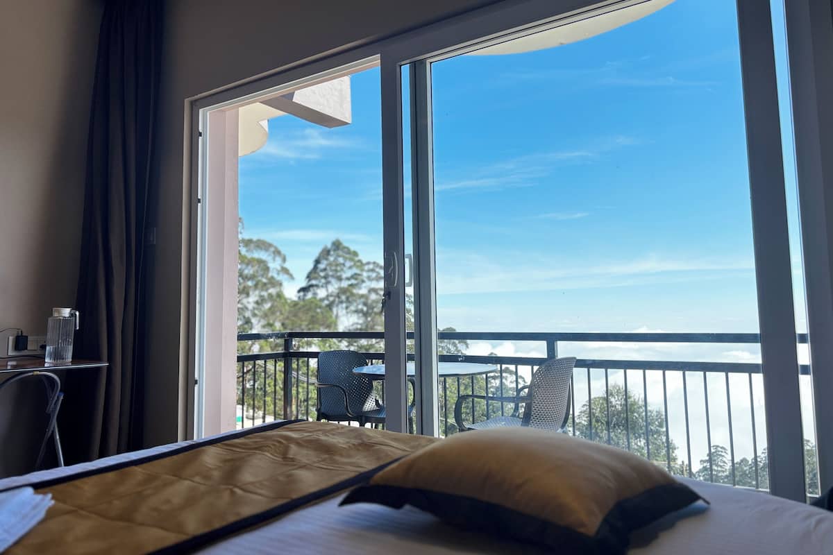 A hotel room with a large window, a bed with a pillow, a chair, and a table with a glass of water.