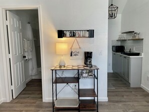 A hallway with a table, a hanging sign, a lamp, and a kitchen area with cabinets and a microwave.