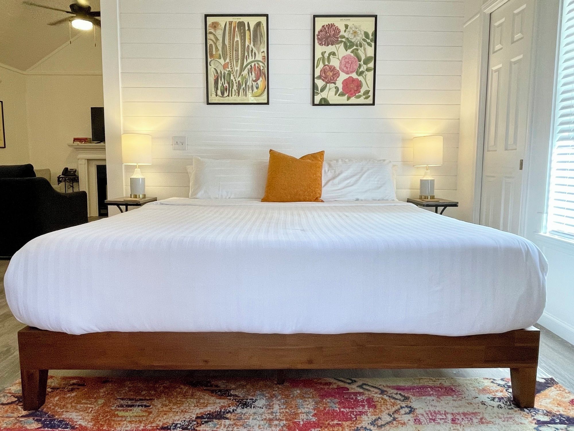 A large bed with white bedding, a wooden frame, and an orange accent pillow. There are two wall-mounted lamps and two framed botanical prints above the bed.