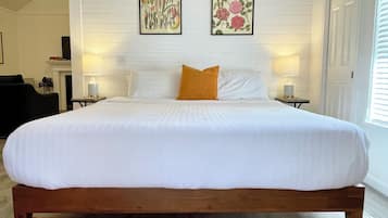 A large bed with white bedding, a wooden frame, and an orange accent pillow. There are two wall-mounted lamps and two framed botanical prints above the bed.