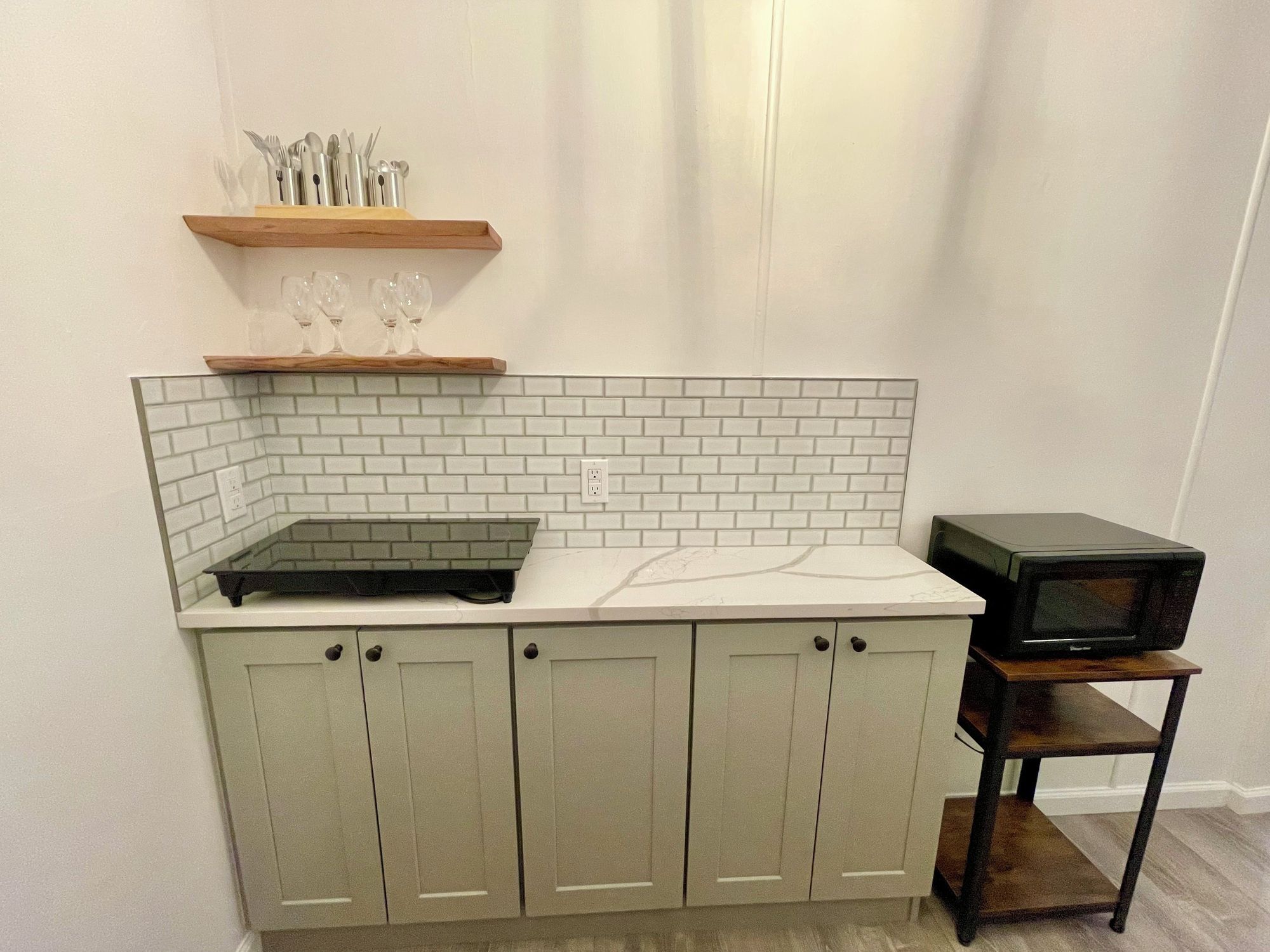 A kitchen with white subway tiles, a microwave on a wooden shelf, and a shelf with cutlery and glasses.