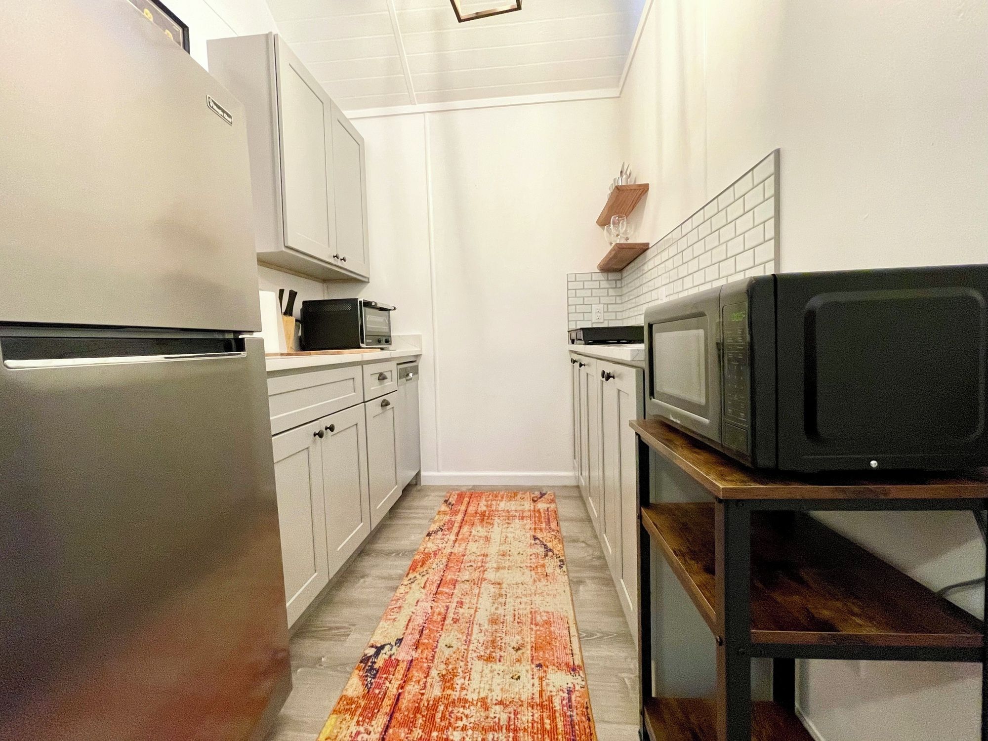 A compact kitchen with white cabinets, a refrigerator, a microwave, and a wooden shelf.