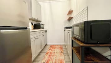 A compact kitchen with white cabinets, a refrigerator, a microwave, and a wooden shelf.