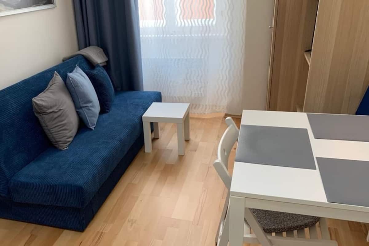 A modern living room with a blue sofa, a small white table, and a dining area with a white table and chairs.