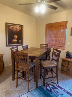 A dining area with a wooden table and chairs, a ceiling fan, a window with blinds, and a painting on the wall.