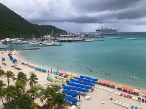 Una playa con hileras de sombrillas y tumbonas, un puerto deportivo con barcos, y un crucero amarrado al fondo.