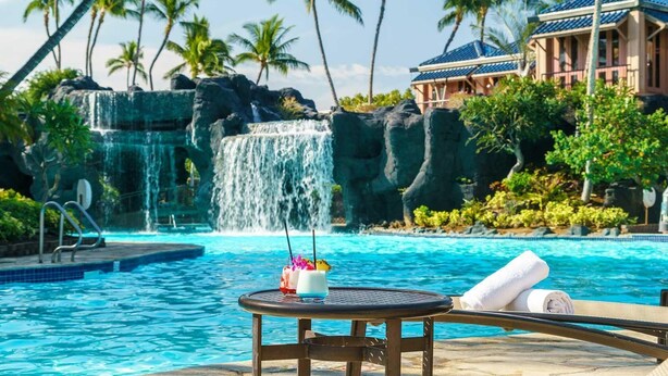 A pool with a waterfall, palm trees, and a lounge area with a table and chairs. - Fairway Villas Waikoloa G31 (Waikoloa)